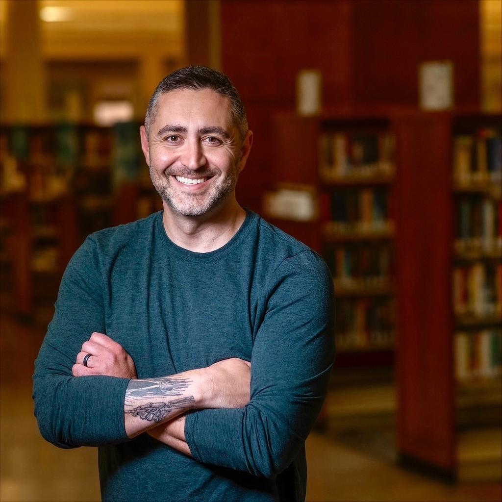 Portrait of Dmitri Atrash smiling with arms crossed in a library